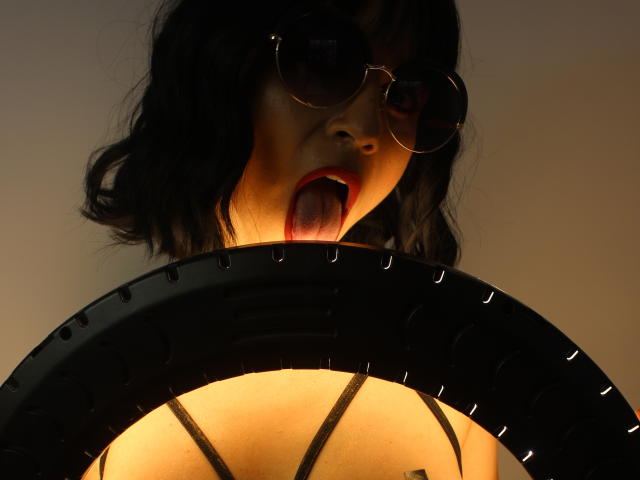 SexyKonnyX met donker krullend haar, die ronde zonnebril draagt, haar rode tong uitsteekt, omhoog kijkt, en omringd is door een zwart, cirkelvormig object. Ze draagt een zwarte top met spaghettibandjes, de belichting is warm, en het is een foto..