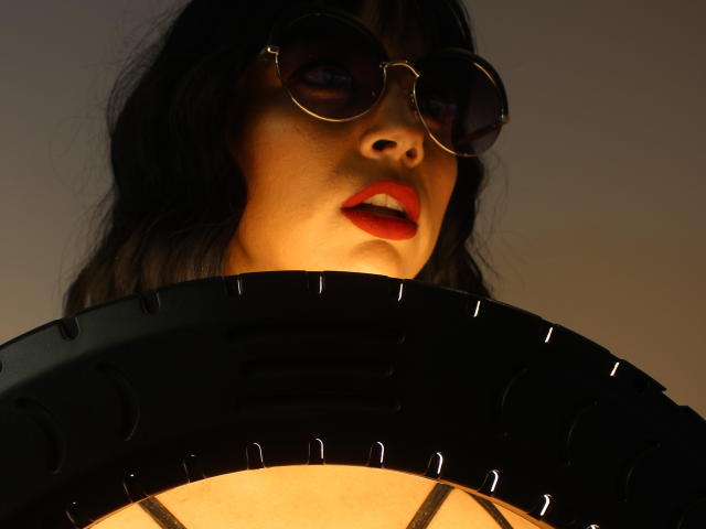SexyKonnyX mit dunklem Haar, roten Lippen, die runde Sonnenbrille trägt und im Vordergrund eine schwarze, runde Lampe mit warmem Schein zu sehen ist, blickt nach unten und hat den Mund leicht geöffnet..