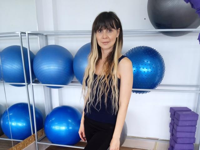 LovenaHelen, con cabello largo y degradado de negro a rubio, vestida con una camiseta azul marino sin mangas y pantalones negros, está de pie en una habitación con pelotas de ejercicio azules y moradas..