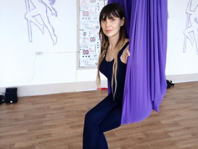 LovenaHelen, com longos cabelos castanhos, vestindo um vestido azul justo, está sentada parcialmente escondida por cortinas roxas em um quarto com piso de madeira, exibindo seus pequenos seios e traseiro esguio..