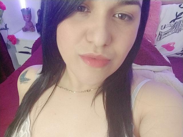 VivianaThompson has pale skin, dark straight hair, and glossy pink lips. She's wearing a white tank top, showing small tits and a butterfly tattoo on her left shoulder.