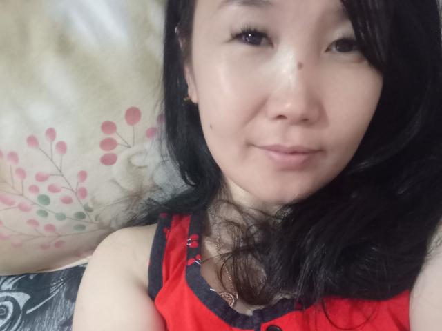 BetLu has light skin, black hair, and a slight smile. She's wearing a red lace top, showing her small, cute tits. She's lying down, looking at the camera.