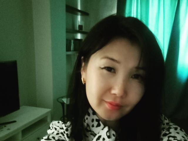BetLu with black hair, light skin, and red lipstick, smiling softly, wearing a black-and-white patterned top, and gold earrings, in a dimly lit room with green curtains.