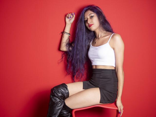 Julia, con cabello largo y morado, top blanco, falda negra y botas negras hasta el muslo, sentada en una silla roja, con una mano levantada, lápiz labial rojo y una pulsera negra..