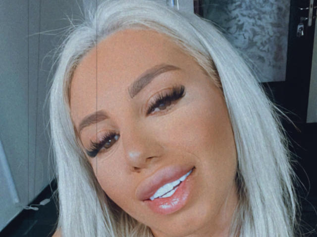 LisaaKiraa has long white hair, dark eyebrows, long eyelashes, white teeth, and glossy pink lips. Her tan skin looks smooth. She's smiling slightly, with a cute, sexy look.