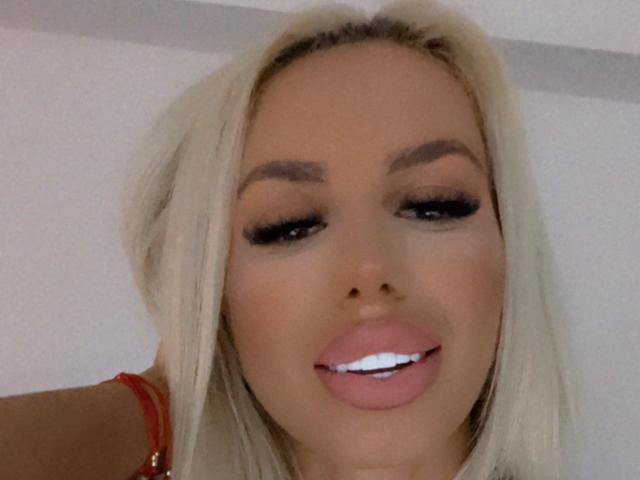 LisaaKiraa a les cheveux blonds, des sourcils foncés, de longs cils, des lèvres roses et brillantes, et elle montre des dents blanches dans un léger sourire. Elle porte une bretelle rouge sur son épaule..