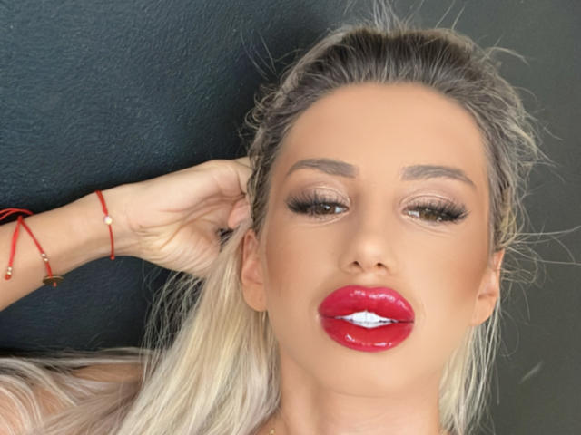 LisaaKiraa has glossy red lips, heavy eyeliner, and blonde hair in a ponytail, wearing a red bracelet against a dark background, with her hand in her hair.