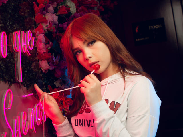 JanneCarter, avec des cheveux roux, un sweat à capuche blanc, suçant un bonbon rouge, tenant un bâton blanc, regardant la caméra, des lettres néon roses à gauche, fond sombre, texte "University of Virginia"..
