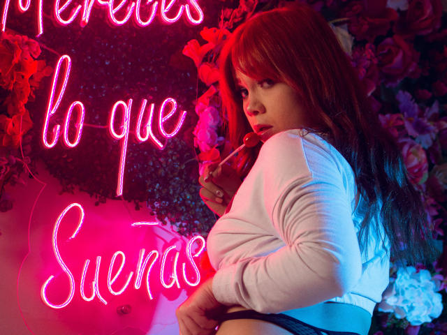 JanneCarter with red hair, white long-sleeve top, holding a lollipop, looking over her shoulder, showing her ass, in a neon pink and blue-lit room.