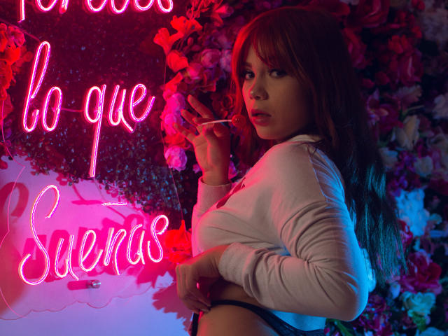 JanneCarter with long brown hair, wearing a white long-sleeve top, holding a cigarette, looking over her shoulder, neon pink light, flower wall, exposed black panties, sexy.