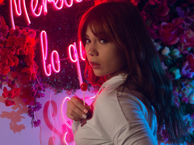 Janne Carter, com cabelos castanhos longos, camisa branca, segurando um pirulito vermelho, luzes rosa neon e rosas vermelhas ao fundo, olhando para trás com uma expressão sedutora..
