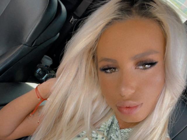 LisaaKiraa with blonde hair, brown eyes, and winged eyeliner, looking down with a slight smile, wearing a red bracelet, in a car.
