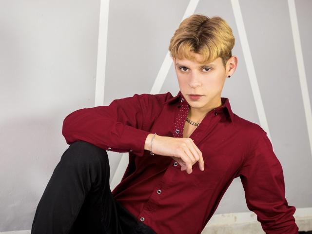 Logan Evans, con cabello rubio corto, vistiendo una camisa a rayas, una bufanda roja, pantalones negros, sentado en una silla roja con tapicería, mirando a la cámara con una expresión seria, con una mano en la rodilla..