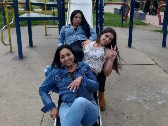 Trois femmes latines lesbiennes dans une aire de jeux, assises sur une glissade. Deux en première ligne, portant des vestes en jean bleu et des jeans, souriant. Une à l'arrière, avec un haut blanc, faisant un signe de la paix..