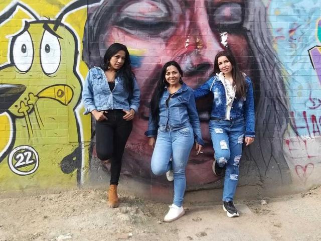 Tres mujeres latinas lesbianas con cabello largo y oscuro, vestidas con jeans azules y chaquetas de mezclilla, de pie frente a un grafiti, sonriendo, y una de ellas levantando una pierna. Una de ellas lleva botas marrones..