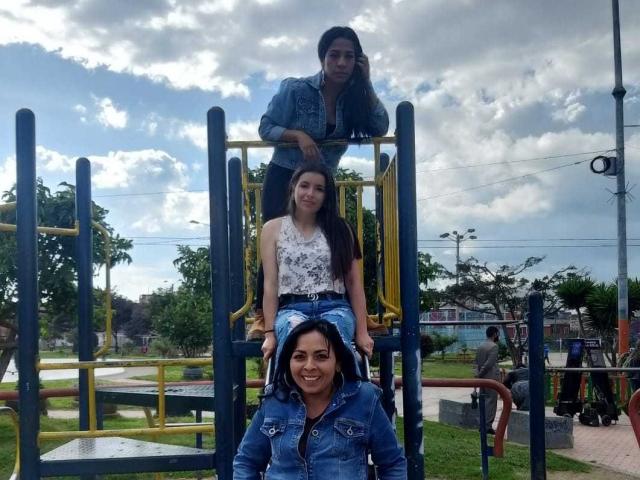 Three LatinLesbians on a playground: Lateinamerikanerin mit langen, schwarzen Haaren, Jeansjacke und großer Brust, lateinamerikanische Frau mittleren Alters mit weißem Oberteil mit Blumenmuster und gutem Hintern, lateinamerikanische Frau unten mit blauer Jacke, lächelnd..