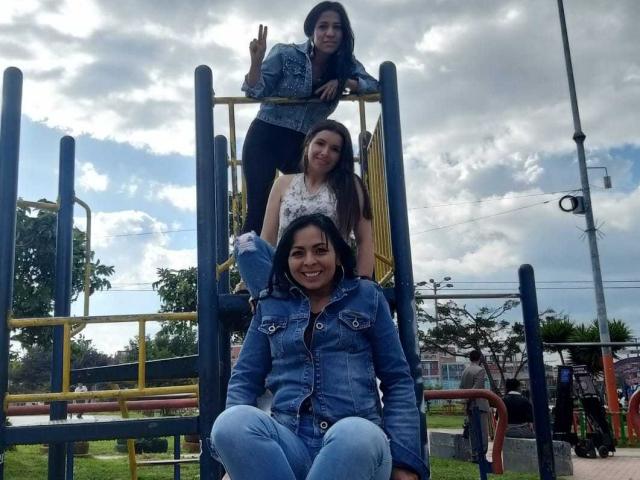 Drei lateinamerikanische Lesben auf einem Spielplatz, die Jeans tragen, stehen und sitzen auf einer gelben Metallkonstruktion. Eine hat kleine Brüste, eine andere mittelgroße Brüste, und das Gesäß einer ist verdeckt..