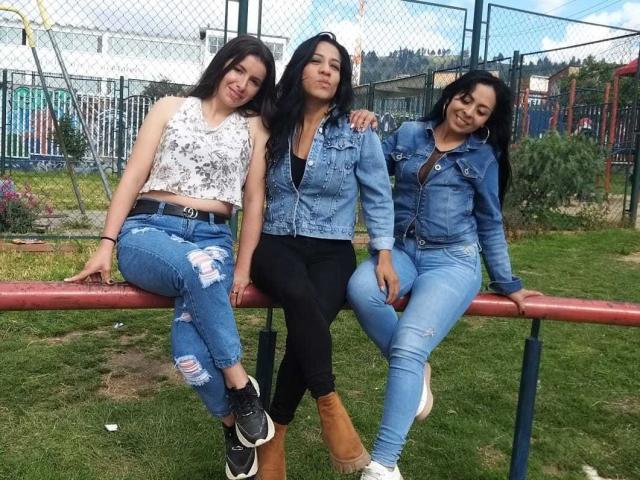 Drie Latina vrouwen, lesbisch, met lang donker haar, die spijkerjassen en versleten jeans dragen, zitten op een rode metalen bank, met hun armen om elkaar heen, lachend, in een grasrijk park..