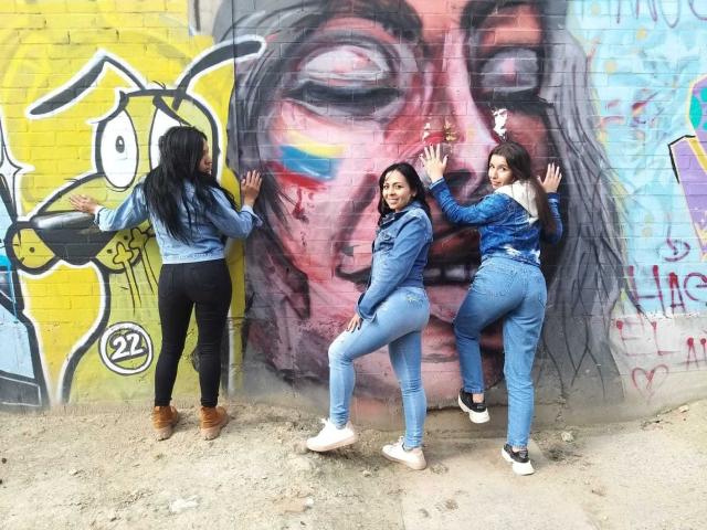 Drie Latina vrouwen, lesbisch, gekleed in blauwe jeans en denim jassen, staan met hun rug tegen een muur en raken kleurrijke graffiti van een gezicht aan. Twee hebben een zichtbare achterkant, één heeft lang zwart haar..