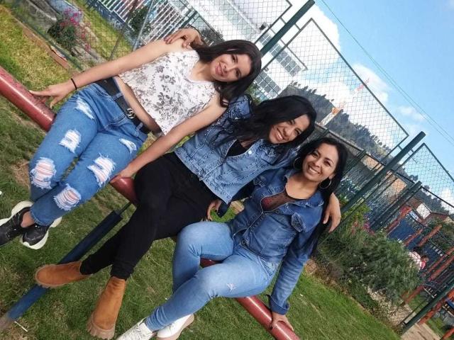 Drie Latina vrouwen, lesbisch, met zwart haar, die spijkerjassen en versleten jeans dragen, zitten op een rode metalen stang en glimlachen. Op de achtergrond gras, een hek en een blauwe lucht..