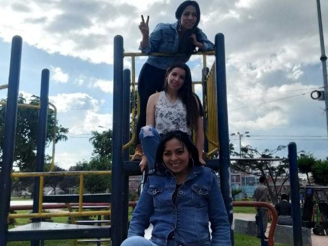Três mulheres latinas em um parquinho, em pé e sentadas em uma estrutura de metal amarela e azul. A mulher latina do topo faz o sinal da paz, a do meio veste um vestido floral branco, e a de baixo usa uma jaqueta jeans..