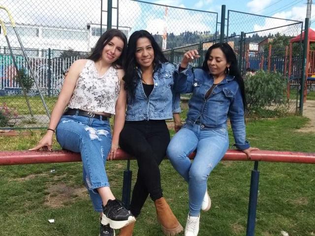 Trois femmes latines, lesbiennes, aux cheveux foncés, portant du denim, assises sur un banc rouge. L'une porte un haut blanc, l'autre un pantalon noir, et la troisième un jean bleu. Toutes sourient, les bras enlacés..
