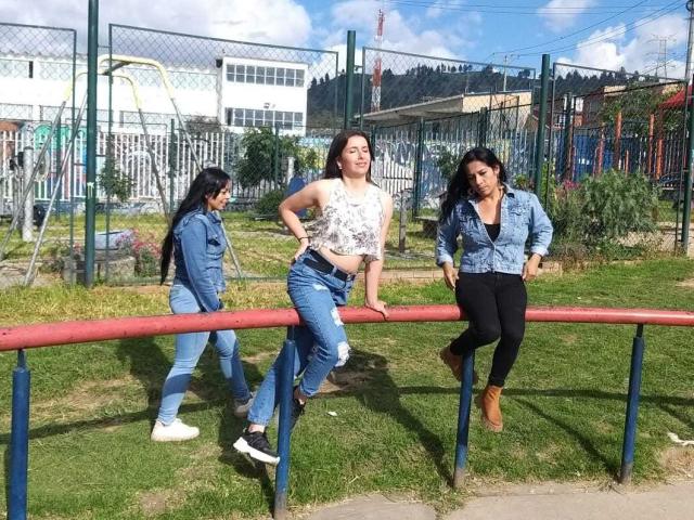 Drie Latina's die leunen op een rode metalen stang in een park. De Latina in het midden heeft grote borsten, draagt een witte crop top en gescheurde jeans. De Latina's links en rechts dragen denim jassen..