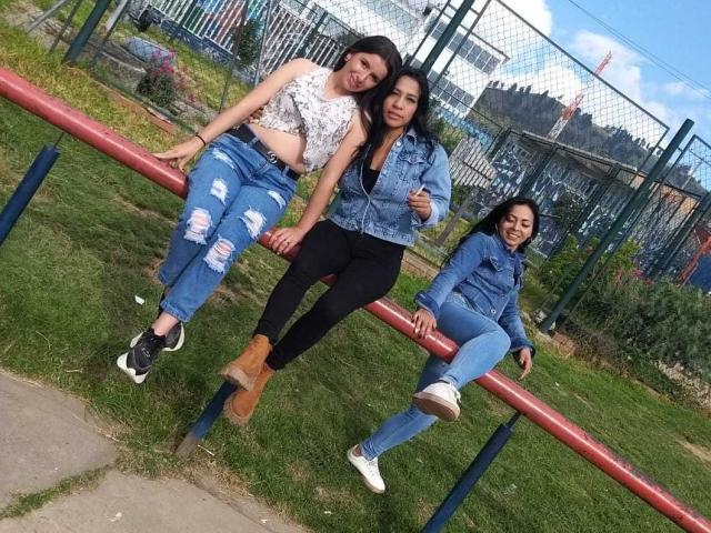 Drei lateinamerikanische, lesbische Frauen lehnen sich an eine rote Metallreling, tragen Jeansjacken und zerrissene blaue Jeans, braune Stiefel und weiße Turnschuhe und lächeln vor der Kamera im Freien..