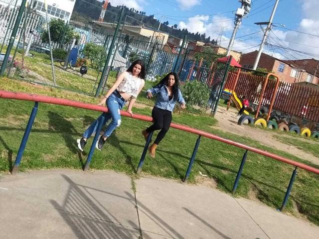 Een LatinLesbian met borsten, gekleed in een witte top, blauwe jeans en laarzen, en een andere LatinLesbian in een blauw shirt en zwarte broek, die aan een rode reling in een park hangt..