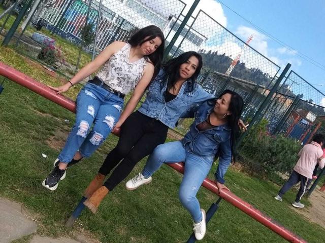 Tre donne latine lesbiche con capelli scuri, che indossano giacche di jeans e pantaloni strappati, sedute su una recinzione di metallo rossa in un'area erbosa. La donna latina lesbica a sinistra ha piccoli seni, quella al centro ha i glutei prominenti..