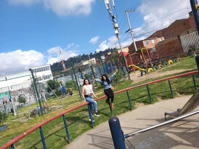 Een Latina lesbische vrouw met borsten en een ronding, gekleed in een wit shirt en blauwe jeans, zit op een rode hek. Een andere Latina lesbische vrouw, in een blauw shirt en zwarte rok, springt naast haar..