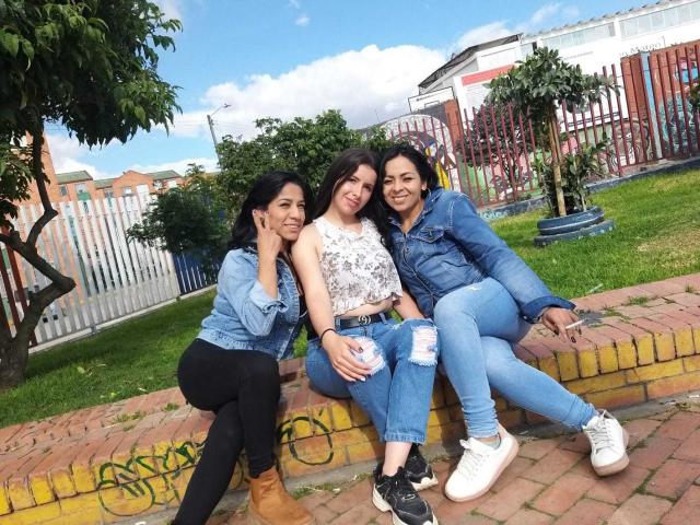 Três mulheres latinas, lésbicas, sentadas em um muro de tijolos, sorrindo, vestindo jaquetas de jeans e calças jeans rasgadas, com botas pretas e brancas, em um quintal ensolarado, com grama verde e céu azul..