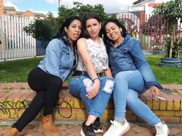 Três mulheres latinas lésbicas sentadas em um muro de tijolos. A mulher latina lésbica à esquerda tem cabelo preto, colete azul e calças pretas. A mulher latina lésbica do meio tem cabelo castanho, blusa branca e jeans rasgados. A mulher latina lésbica à direita tem cabelo preto, colete azul e jeans azuis..