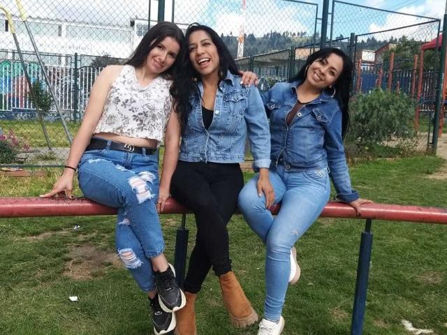 Três mulheres latinas, com cabelos longos e pretos, vestindo jaquetas de jeans e calças jeans rasgadas, sentadas em um balcão vermelho, sorrindo, mostrando seus seios e nádegas em um parque gramado..