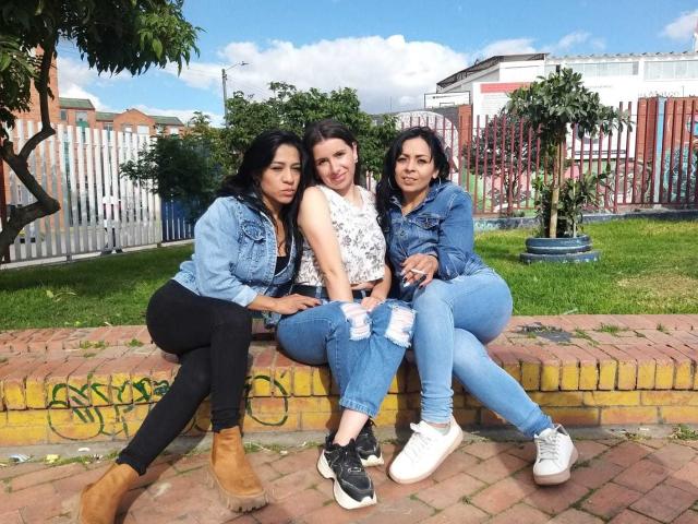 Three LatinLesbians sitting on a brick wall, wearing denim jackets and jeans, with black, white, and blue tops, and tan, black, and white sneakers, under a clear blue sky.