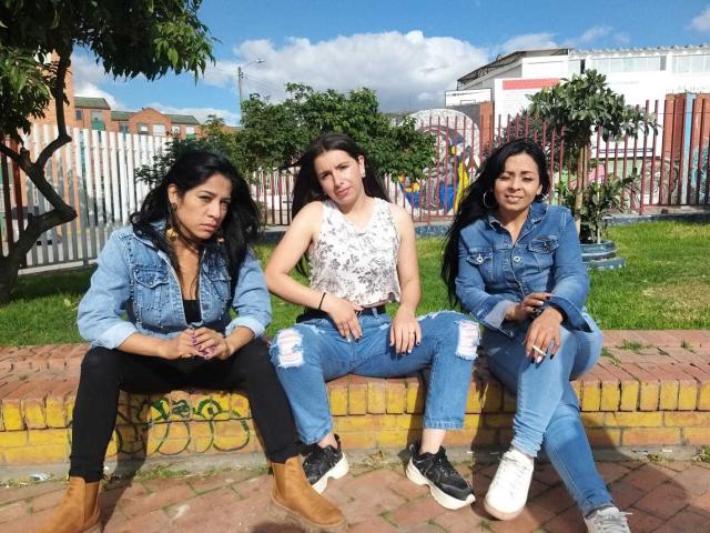 Tre donne lesbiche di origine latina, con capelli neri, che indossano giacche di jeans e pantaloni strappati, sedute su un muro di mattoni, che mostrano i loro glutei con scarpe da ginnastica nere e bianche, sotto un cielo azzurro e soleggiato..