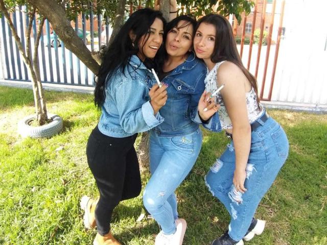 Drie Latina vrouwen die lesbisch zijn, met lang zwart haar, gekleed in denim jassen en versleten jeans, staan op het gras, glimlachen en leunen tegen een boom, terwijl ze ijsjes vasthouden..