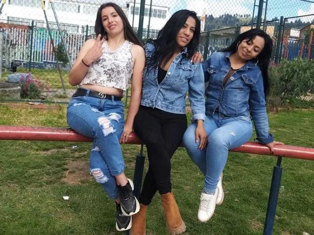Tre donne latine lesbiche sedute su una recinzione rossa, che indossano giacche di jeans e pantaloni strappati. Quella a sinistra indossa una canotta bianca, quella al centro una maglietta nera, e quella a destra una giacca nera..