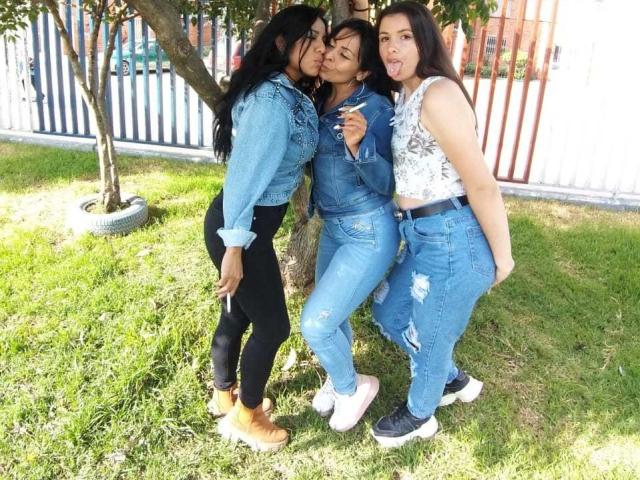 Três mulheres latinas, abertamente lésbicas, estão em um gramado, se beijando e posando. Elas vestem calças e jaquetas de jeans azuis, blusas pretas e brancas, com botas e tênis..
