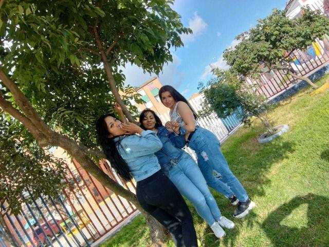 Trois femmes lesbiennes d'origine latine debout dans une cour verdoyante, portant des vestes en jean et des jeans, avec des arbres et une clôture en arrière-plan. L'une d'elles fait un signe de la paix et sourit..