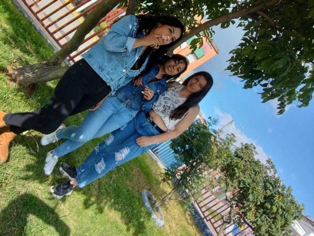 Três mulheres latinas, lésbicas, em pé na grama, vestindo jeans e jaquetas de sarja, uma com calças pretas e botas marrons, todas sorrindo e mostrando sinais de paz, sob uma árvore..