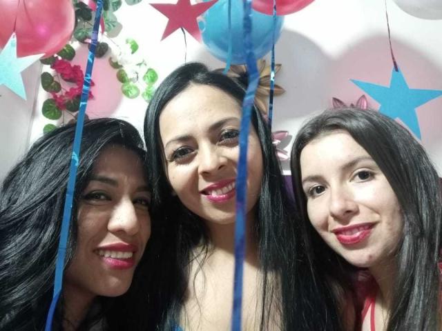 Drei lateinamerikanische, lesbische Frauen mit langen, schwarzen Haaren, rosa Lippenstift und lächelnden Gesichtern, die eng beieinander stehen, umgeben von blauen und roten, sternförmigen Ballons und rosa, grünen und silbernen Girlanden..