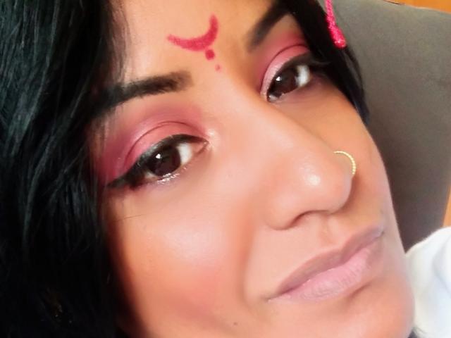 Nahirr has pink eyeshadow, dark eyebrows, a red forehead dot, black hair, a gold nose ring, and a slight smile. Her skin is tan and she's looking at the camera.