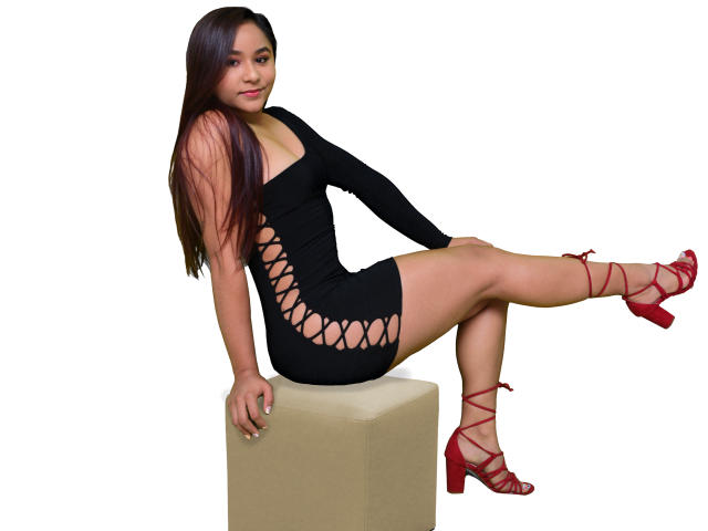 JessicaFit sentada sobre un cubo, vistiendo un vestido negro con detalles recortados a los lados, tacones rojos con tiras y cabello largo castaño. Sus piernas están cruzadas, mostrando su trasero..
