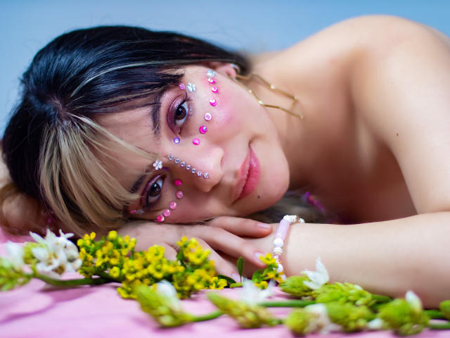 ElizabethBlizz con segni sul viso a forma di gemma rosa, capelli biondi e neri, sdraiata senza top, braccialetto rosa, fiori gialli, petali bianchi e foglie verdi su una superficie rosa, che guarda verso la fotocamera..
