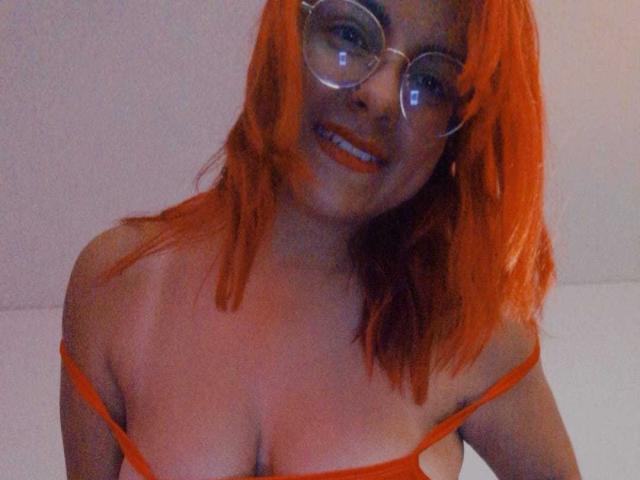 EmmaLuna, com cabelo laranja, óculos e lingerie laranja, mostrando os seios, sorrindo com batom laranja, em um quarto com pouca luz..