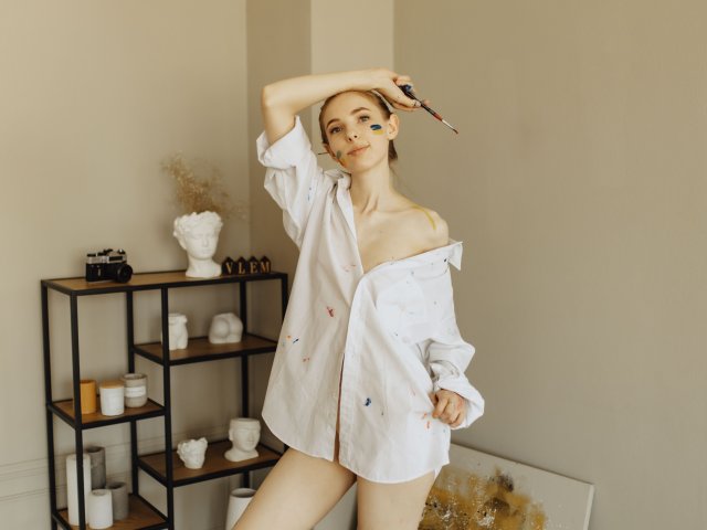 SafiraFuracao con maquillaje azul en los ojos, vistiendo una camisa blanca con un hombro descubierto, sosteniendo un pincel, en una habitación minimalista con estanterías decoradas en blanco y negro. Sus senos son visibles..