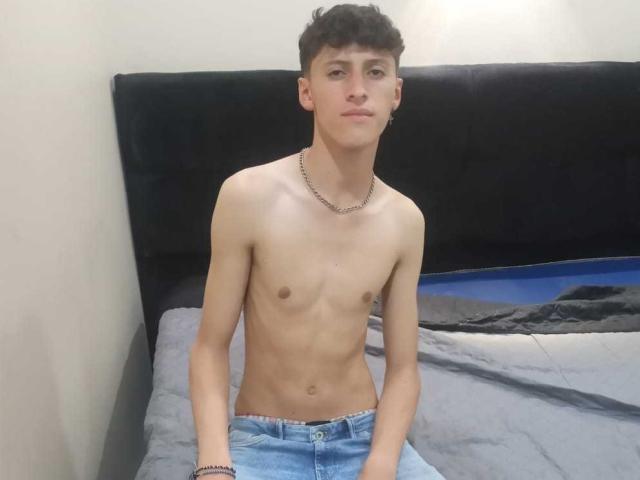 Carlos Rubiio, senza camicia, magro con piccoli seni, indossa jeans azzurri chiari, una collana d'argento, è seduto sul letto, ha i capelli castani ricci e corti, è leggermente attraente, con un'espressione neutra, ha la pelle chiara, aspetto casual..