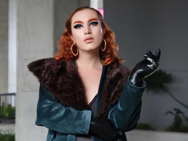 JuliaFanning with orange curly hair, wearing a green velvet coat with dark fur, black gloves, and gold hoop earrings, making a "shush" gesture with her finger to her lips.
