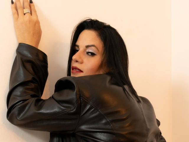 LizaRussell with black leather jacket, black nails, ring on finger, looking over shoulder, red lipstick, black hair, against white wall, hand on wall, sexy, sultry expression.